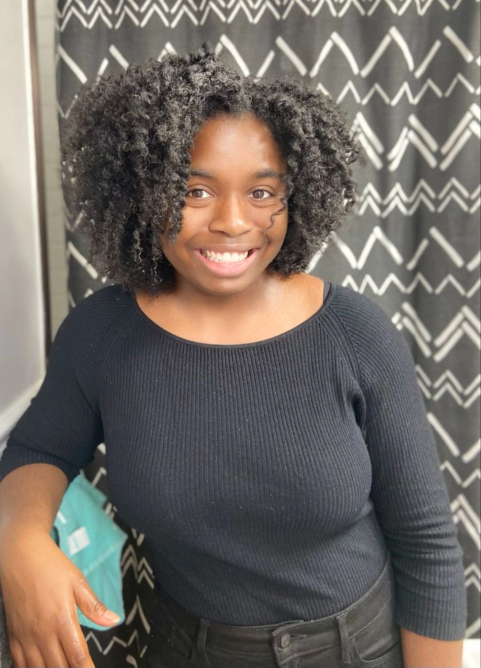 A woman with a neat, medium-length twist out hairstyle in an office setting, wearing a professional blazer and smiling slightly